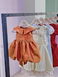 A row of children's dresses is hanging on a metal clothing rack. The dresses are on wooden hangers that are labeled 'Gisou'. The garments feature various colors and styles, with a prominent orange dress in the front that has a classic, flared skirt and a simple collar. Other dresses in lighter shades, as well as a vibrant red dress, are visible in the background.