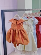 A row of children's dresses is hanging on a metal clothing rack. The dresses are on wooden hangers that are labeled 'Gisou'. The garments feature various colors and styles, with a prominent orange dress in the front that has a classic, flared skirt and a simple collar. Other dresses in lighter shades, as well as a vibrant red dress, are visible in the background.