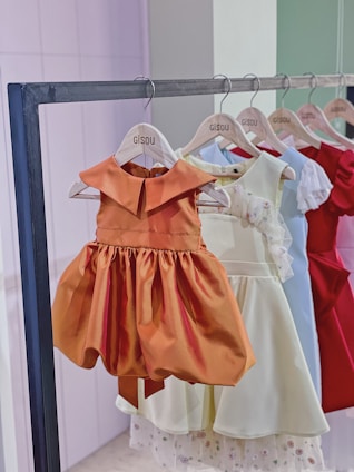 A row of children's dresses is hanging on a metal clothing rack. The dresses are on wooden hangers that are labeled 'Gisou'. The garments feature various colors and styles, with a prominent orange dress in the front that has a classic, flared skirt and a simple collar. Other dresses in lighter shades, as well as a vibrant red dress, are visible in the background.