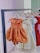 A row of children's dresses is hanging on a metal clothing rack. The dresses are on wooden hangers that are labeled 'Gisou'. The garments feature various colors and styles, with a prominent orange dress in the front that has a classic, flared skirt and a simple collar. Other dresses in lighter shades, as well as a vibrant red dress, are visible in the background.