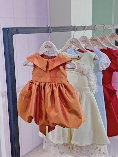 A row of children's dresses is hanging on a metal clothing rack. The dresses are on wooden hangers that are labeled 'Gisou'. The garments feature various colors and styles, with a prominent orange dress in the front that has a classic, flared skirt and a simple collar. Other dresses in lighter shades, as well as a vibrant red dress, are visible in the background.