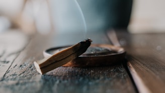 A wooden incense stick is burning on a wooden surface, with smoke gently rising from its charred end. The focus is on the stick and its holder, while the background is softly blurred.