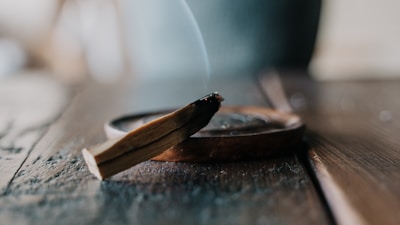 A wooden incense stick is burning on a wooden surface, with smoke gently rising from its charred end. The focus is on the stick and its holder, while the background is softly blurred.