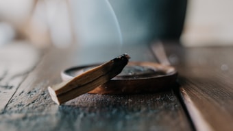 A wooden incense stick is burning on a wooden surface, with smoke gently rising from its charred end. The focus is on the stick and its holder, while the background is softly blurred.