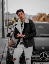 A man is leaning against a black Mercedes car on a city street. He is wearing a stylish black leather jacket with a white fur collar, a patterned shirt, white ripped jeans, and a watch. The background shows an urban setting with trees and buildings, suggesting a chilly autumn day.