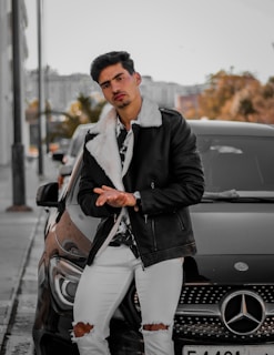 A man is leaning against a black Mercedes car on a city street. He is wearing a stylish black leather jacket with a white fur collar, a patterned shirt, white ripped jeans, and a watch. The background shows an urban setting with trees and buildings, suggesting a chilly autumn day.