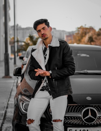 A man is leaning against a black Mercedes car on a city street. He is wearing a stylish black leather jacket with a white fur collar, a patterned shirt, white ripped jeans, and a watch. The background shows an urban setting with trees and buildings, suggesting a chilly autumn day.