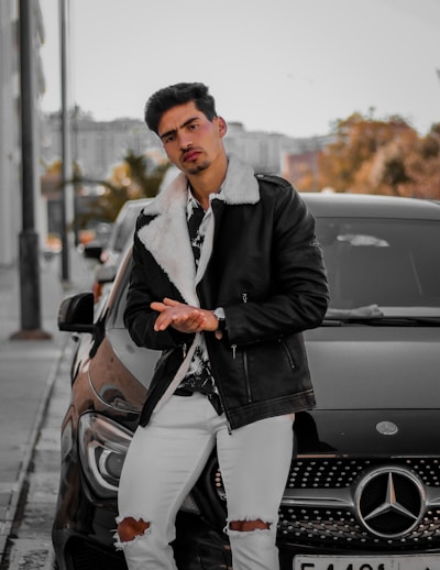 A man is leaning against a black Mercedes car on a city street. He is wearing a stylish black leather jacket with a white fur collar, a patterned shirt, white ripped jeans, and a watch. The background shows an urban setting with trees and buildings, suggesting a chilly autumn day.