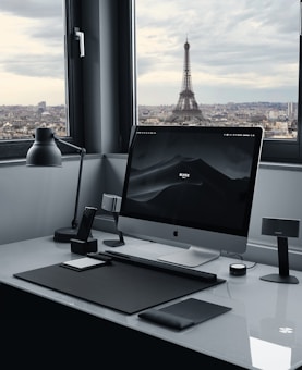 A modern workspace with a large computer monitor on a sleek white desk. Various desk accessories, including a lamp, speakers, and a smartphone stand, are neatly arranged. Through the large window, the Eiffel Tower is visible against a cloudy sky, offering a scenic city view.