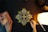 An intricate geometric mandala design is being drawn with gold ink on a black paper. The image includes two hands, one holding a pen and the other resting on the table, adorned with rings. There is a glowing lamp on the right side, casting warm light, and a book titled 'The Secret' on the left.