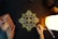 An intricate geometric mandala design is being drawn with gold ink on a black paper. The image includes two hands, one holding a pen and the other resting on the table, adorned with rings. There is a glowing lamp on the right side, casting warm light, and a book titled 'The Secret' on the left.
