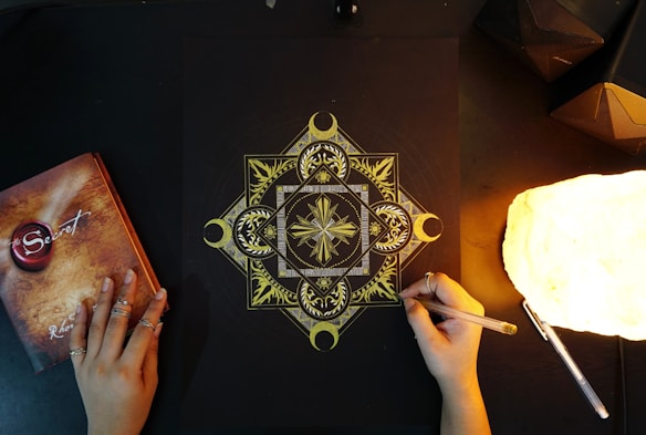 An intricate geometric mandala design is being drawn with gold ink on a black paper. The image includes two hands, one holding a pen and the other resting on the table, adorned with rings. There is a glowing lamp on the right side, casting warm light, and a book titled 'The Secret' on the left.