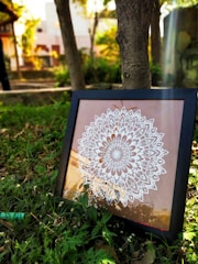 A framed artwork with a detailed white mandala pattern placed on the grass in an outdoor setting. Sunlight filters through the leaves, casting shadows and creating light streaks on the print. The background includes trees and blurred structures, suggesting a garden or park environment.