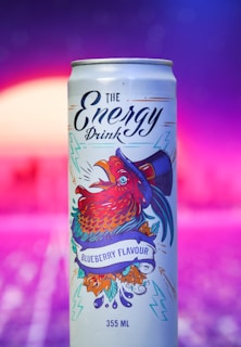 A can of blueberry-flavored energy drink featuring vibrant artwork of a rooster wearing a hat. The can design includes colorful illustrations with electric blue and red hues, surrounded by decorative elements like leaves and lightning bolts. The background is a gradient of purples and pinks with a soft, blurred effect.