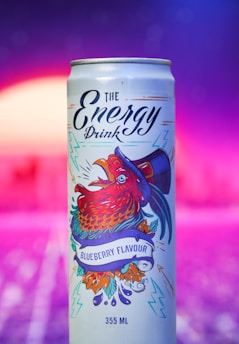 A can of blueberry-flavored energy drink featuring vibrant artwork of a rooster wearing a hat. The can design includes colorful illustrations with electric blue and red hues, surrounded by decorative elements like leaves and lightning bolts. The background is a gradient of purples and pinks with a soft, blurred effect.