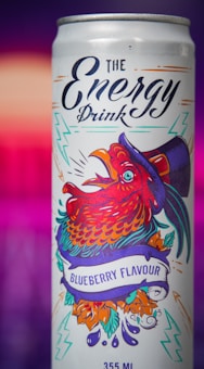 A vibrant energy drink can featuring a stylized red rooster wearing a top hat. The can is decorated with colorful graphics, including lightning bolts and a banner reading 'Blueberry Flavour.' The background is a gradient of pink and purple hues.
