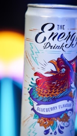 A can of energy drink with vibrant artwork featuring a stylized rooster in dynamic colors. The rooster is surrounded by artistic lightning bolts and a banner indicating a blueberry flavor. The design is vivid, with a contrast between the colorful illustration and the plain background.