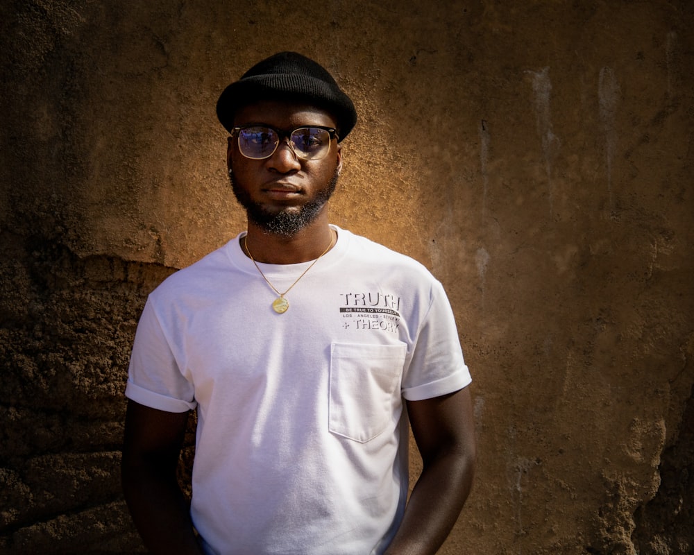 a man wearing a hat and sunglasses standing in front of a stone wall