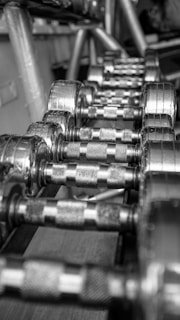 A row of shiny metal dumbbells arranged neatly on a rack. The surface of the dumbbells features a textured grip for enhanced handling. The perspective is from a close-up angle, emphasizing the repetition and alignment of the weights.