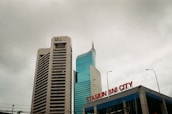 Two tall buildings rise against a cloudy sky. The building on the left is labeled with a BNI logo, featuring a modern design with horizontal lines. The building on the right has a sleek, curved design with reflective glass windows. In the foreground, a sign reads 'STASIUN BNI CITY' in red letters.