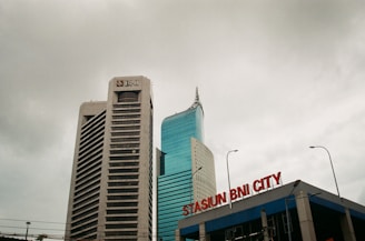 Two tall buildings rise against a cloudy sky. The building on the left is labeled with a BNI logo, featuring a modern design with horizontal lines. The building on the right has a sleek, curved design with reflective glass windows. In the foreground, a sign reads 'STASIUN BNI CITY' in red letters.