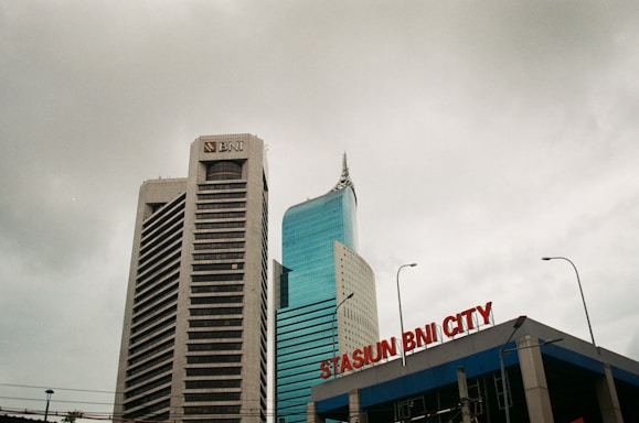 Two tall buildings rise against a cloudy sky. The building on the left is labeled with a BNI logo, featuring a modern design with horizontal lines. The building on the right has a sleek, curved design with reflective glass windows. In the foreground, a sign reads 'STASIUN BNI CITY' in red letters.