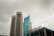 Two tall buildings rise against a cloudy sky. The building on the left is labeled with a BNI logo, featuring a modern design with horizontal lines. The building on the right has a sleek, curved design with reflective glass windows. In the foreground, a sign reads 'STASIUN BNI CITY' in red letters.