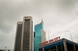 Two tall buildings rise against a cloudy sky. The building on the left is labeled with a BNI logo, featuring a modern design with horizontal lines. The building on the right has a sleek, curved design with reflective glass windows. In the foreground, a sign reads 'STASIUN BNI CITY' in red letters.