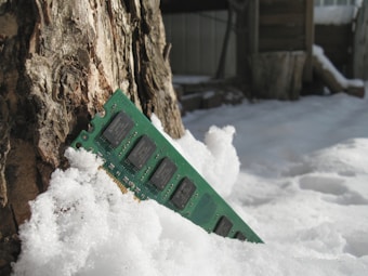 A computer memory module partially buried in snow, resting against the textured trunk of a tree. Sunlight illuminates the snow, creating a bright and somewhat reflective appearance.