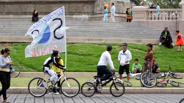 A group of people are gathered around a public square. Several individuals are riding bicycles, one of which has a large flag with a design that includes a flame and water drop. Others are casually standing or sitting on the grass and stone steps. The scene conveys a relaxed and communal atmosphere.