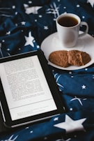 A cozy scene featuring an open ebook reader displaying text, placed on a bed or blanket with a starry pattern. Beside the ebook, there's a white cup filled with a dark beverage, likely coffee or tea, resting on a saucer. Two cookies are placed on the saucer next to the cup.