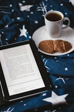 A cozy scene featuring an open ebook reader displaying text, placed on a bed or blanket with a starry pattern. Beside the ebook, there's a white cup filled with a dark beverage, likely coffee or tea, resting on a saucer. Two cookies are placed on the saucer next to the cup.