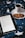 A cozy scene featuring an open ebook reader displaying text, placed on a bed or blanket with a starry pattern. Beside the ebook, there's a white cup filled with a dark beverage, likely coffee or tea, resting on a saucer. Two cookies are placed on the saucer next to the cup.
