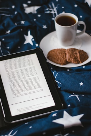 A cozy scene featuring an open ebook reader displaying text, placed on a bed or blanket with a starry pattern. Beside the ebook, there's a white cup filled with a dark beverage, likely coffee or tea, resting on a saucer. Two cookies are placed on the saucer next to the cup.