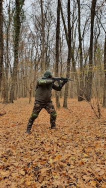 A person dressed in camouflage clothing and protective gear is holding a rifle while standing in a forest. The ground is covered with fallen leaves, and the trees have bare branches typical of autumn.
