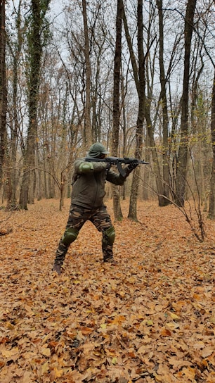 A person dressed in camouflage clothing and protective gear is holding a rifle while standing in a forest. The ground is covered with fallen leaves, and the trees have bare branches typical of autumn.