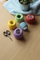 Several colorful yarn balls are arranged on a wooden table next to a pair of scissors. In the background, a potted plant sits on a light blue cloth.