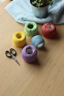 Several colorful yarn balls are arranged on a wooden table next to a pair of scissors. In the background, a potted plant sits on a light blue cloth.