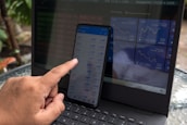 A person is interacting with a smartphone displaying financial data, while a laptop in the background shows various stock market indices. The hand is using a finger to navigate the smartphone. Both screens are filled with numbers and graphs related to market information, with the laptop displaying line charts.