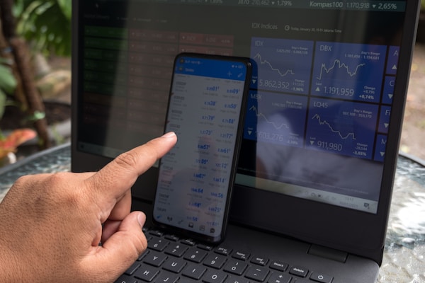 A person is interacting with a smartphone displaying financial data, while a laptop in the background shows various stock market indices. The hand is using a finger to navigate the smartphone. Both screens are filled with numbers and graphs related to market information, with the laptop displaying line charts.