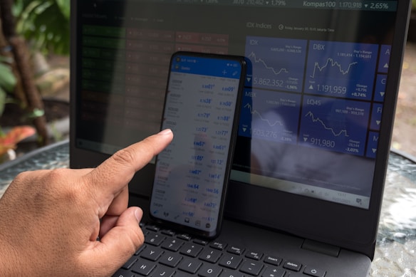 A person is interacting with a smartphone displaying financial data, while a laptop in the background shows various stock market indices. The hand is using a finger to navigate the smartphone. Both screens are filled with numbers and graphs related to market information, with the laptop displaying line charts.