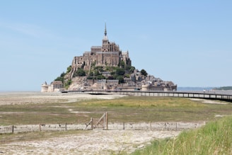 A historic island commune is topped with a grand medieval abbey that rises above its surrounding structures. It is connected to the mainland by a bridge or causeway, with vast open fields and a fence in the foreground. The architecture blends seamlessly with the rocky base, creating a fortress-like appearance.