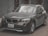 A dark-colored BMW SUV with a slightly dusty exterior is parked on a street. The vehicle has a front license plate from Egypt. The background includes a gray concrete wall and trees with bare branches.