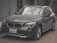 A dark-colored BMW SUV with a slightly dusty exterior is parked on a street. The vehicle has a front license plate from Egypt. The background includes a gray concrete wall and trees with bare branches.