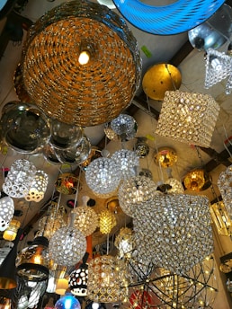 A variety of hanging light fixtures are displayed in a clustered arrangement. The light fixtures include spherical, cylindrical, and cubic designs, many of which are adorned with intricate patterns and reflective surfaces. The colors and styles range from warm golds and silvers to pops of blue.