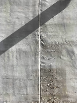 A textured wall with a vertical seam running down the middle. There is a diagonal shadow cast across the surface, adding contrast to the otherwise uniform color. Some imperfections and peeling areas are visible, giving the wall a rugged appearance.