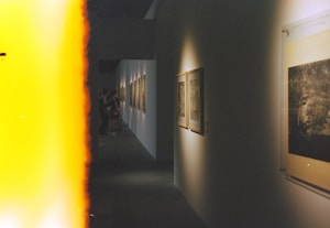 A dimly lit gallery corridor features a series of framed artworks on the walls. The foreground is dominated by a bright yellow light leak from the film, creating an abstract and retro effect. In the background, a few people are standing and observing the art pieces.