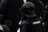 A group of individuals wearing dark, heavy protective gear and helmets. The word 'Полиция' is visible on one of their uniforms, indicating they are police officers. The gear suggests readiness for riot control or similar situations.