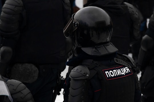A group of individuals wearing dark, heavy protective gear and helmets. The word 'Полиция' is visible on one of their uniforms, indicating they are police officers. The gear suggests readiness for riot control or similar situations.