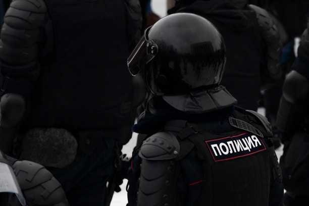 A group of individuals wearing dark, heavy protective gear and helmets. The word 'Полиция' is visible on one of their uniforms, indicating they are police officers. The gear suggests readiness for riot control or similar situations.